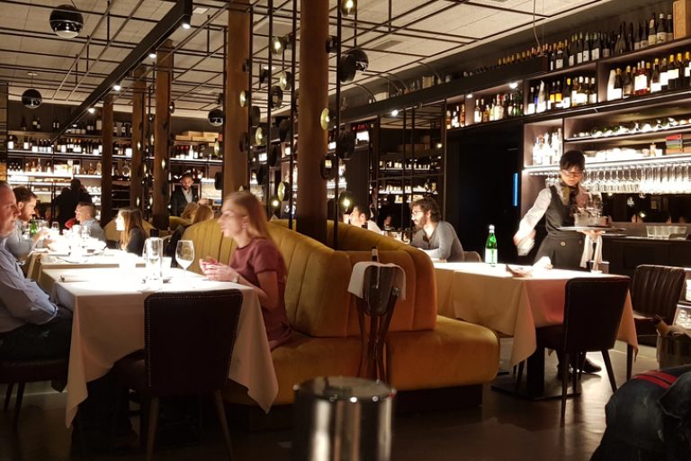 Busy dining room with patrons and staff in a noisy restaurant, highlighting acceptable noise levels in restaurants and ideas on how to reduce noise in a restaurant for better ambience.